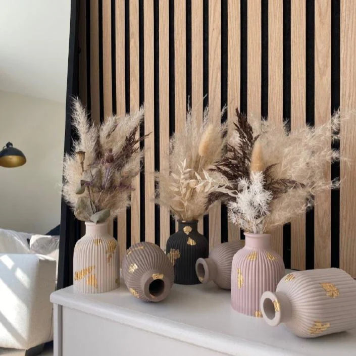 Decorative vases with dried plants on a white surface against a black and wood slat background