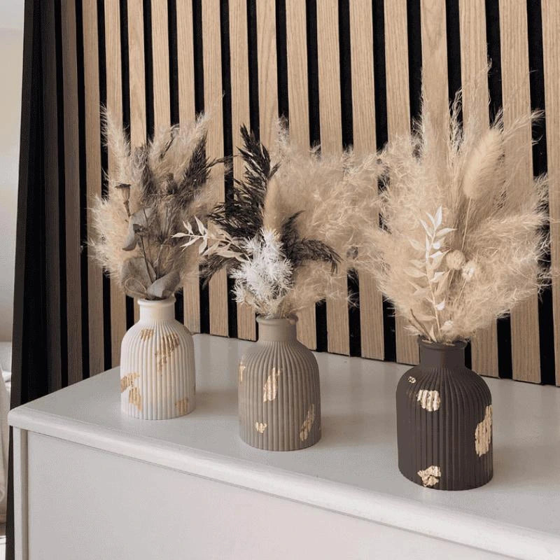 Three decorative vases with dried plants on a white surface against a striped black and beige wall.
